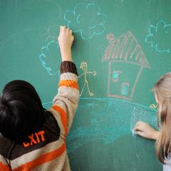 Två barn ritar hus och moln på en grön tavla med färgad krita.